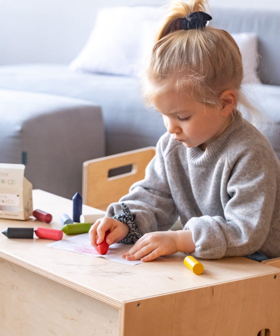 Kindgerecht & bruchfest*Unsere Wachsmalstifte sind stabil und bruchsicher. Ihre ergonomische Form wurde speziell für kleine Kinderhände konzipiert.|Mädchen sitzt zum Malen an Hocker und Bank und malt ein Bild mit den Wachsmalstiften.